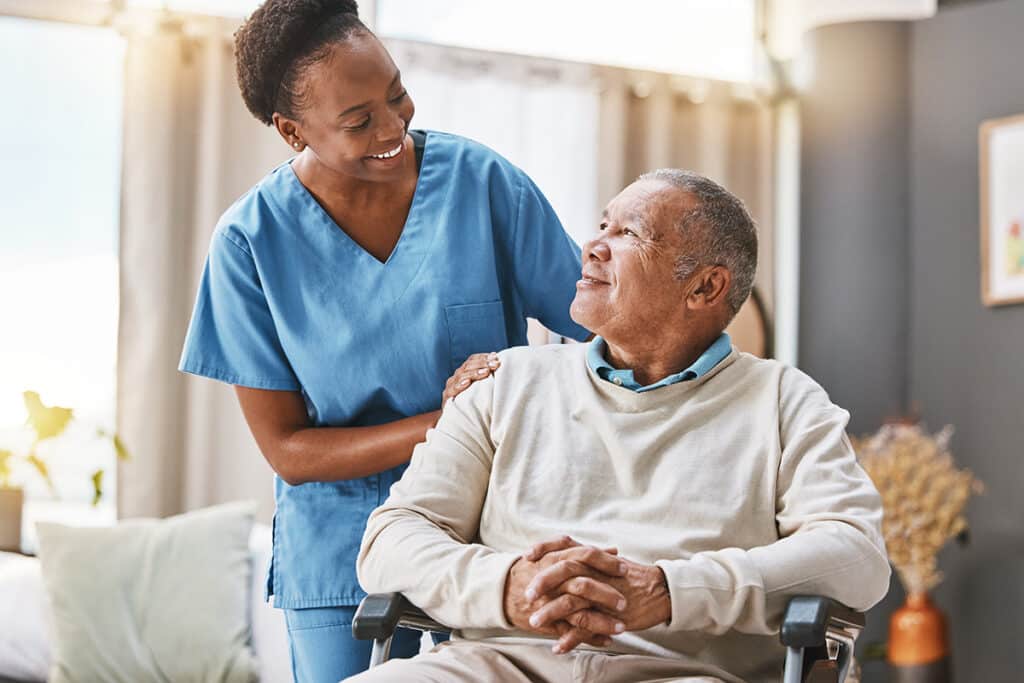 A nurse in blue scrubs smiles warmly at an elderly man in a wheelchair.
