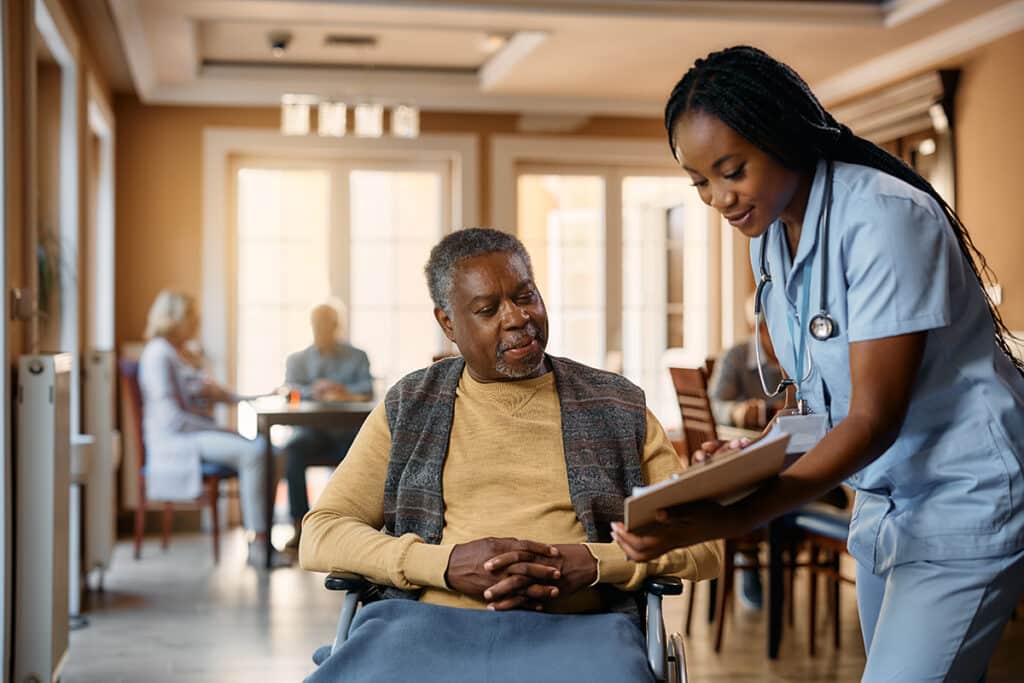 A nurse in blue scrubs smiling and showing a clipboard to a man in a wheelchair