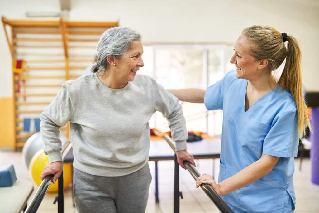 Smiling physiotherapist assisting happy senior woman to walk during movement therapy at rehab center