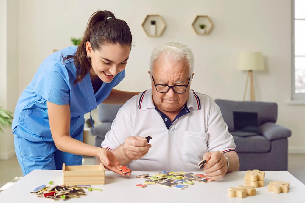 A caregiver in blue scrubs smiles while assisting an elderly man with glasses in assembling a puzzle at a white table, creating an atmosphere of support and care.