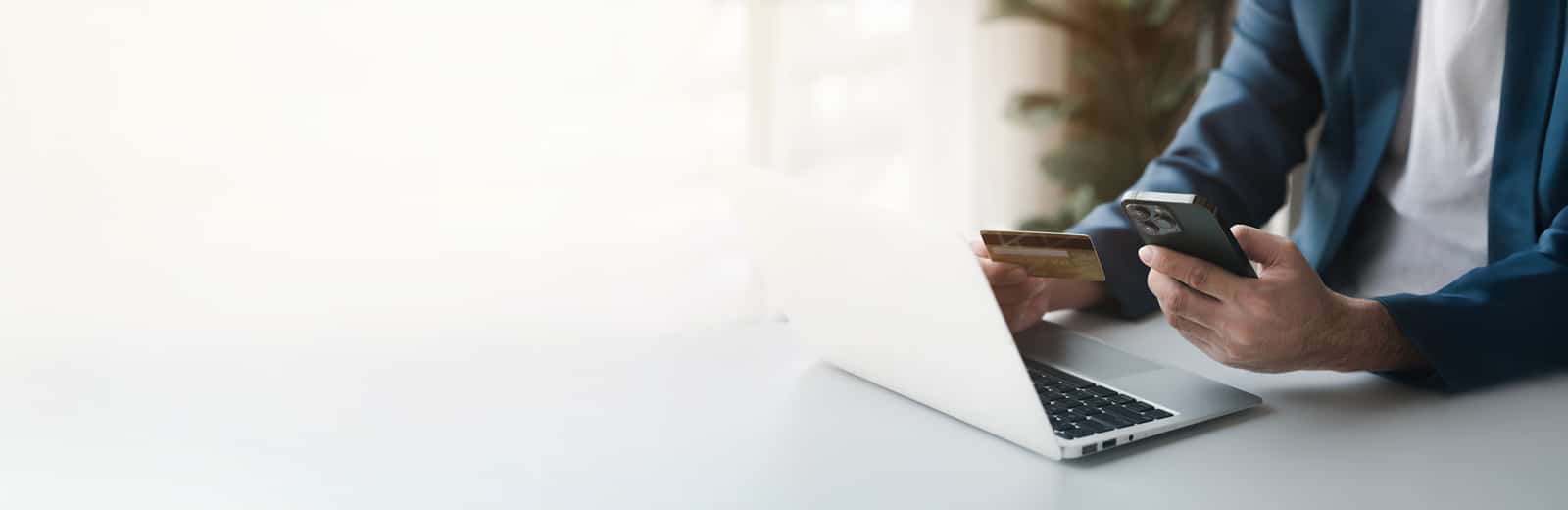 A person in a blue suit is shopping online, holding a credit card and a smartphone, sitting at a white desk with a laptop in a softly lit room