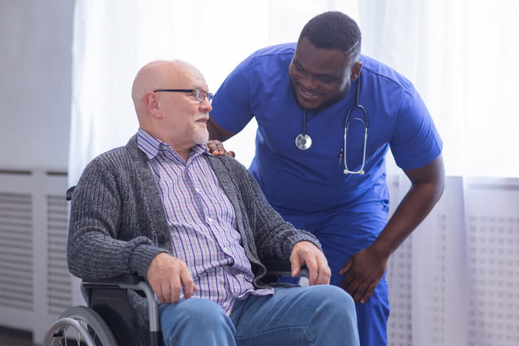 A caring nurse in blue scrubs and stethoscope gently smiles and comforts an elderly man in a wheelchair.