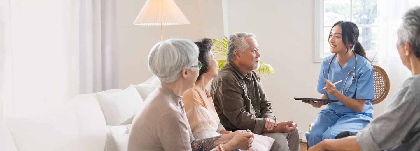 A nurse in blue scrubs holds a tablet, smiling while speaking with a group of seniors seated on a sofa
