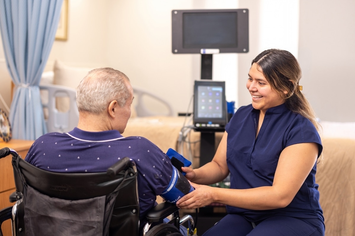 Staff member speaking with a resident seated in a wheelchair beside medical equipment.