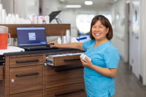 Staff member in blue scrubs smiling beside a laptop at a hallway workstation.