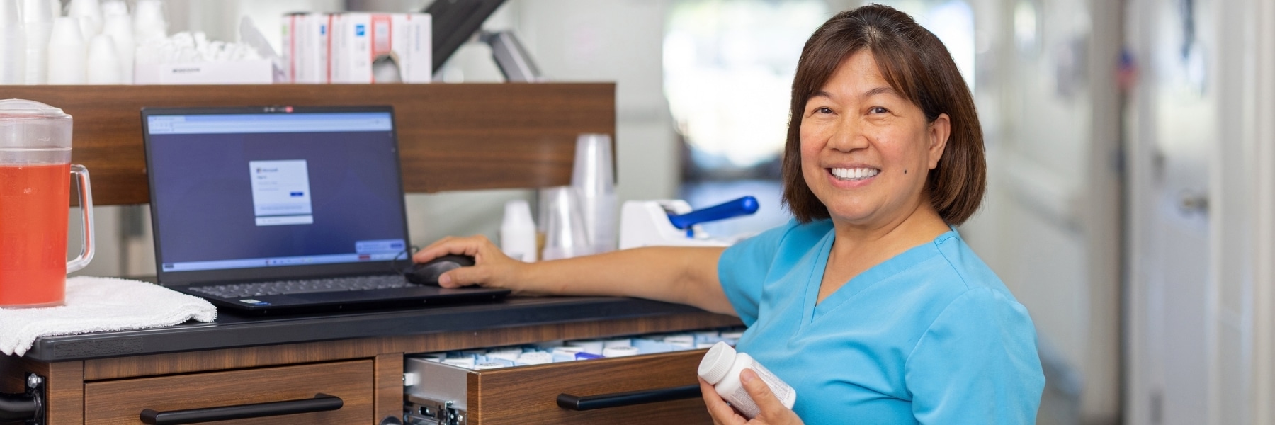 Staff member in blue scrubs smiling beside a laptop and supply cart.
