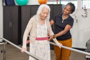Staff member supporting a resident as she walks between parallel bars.