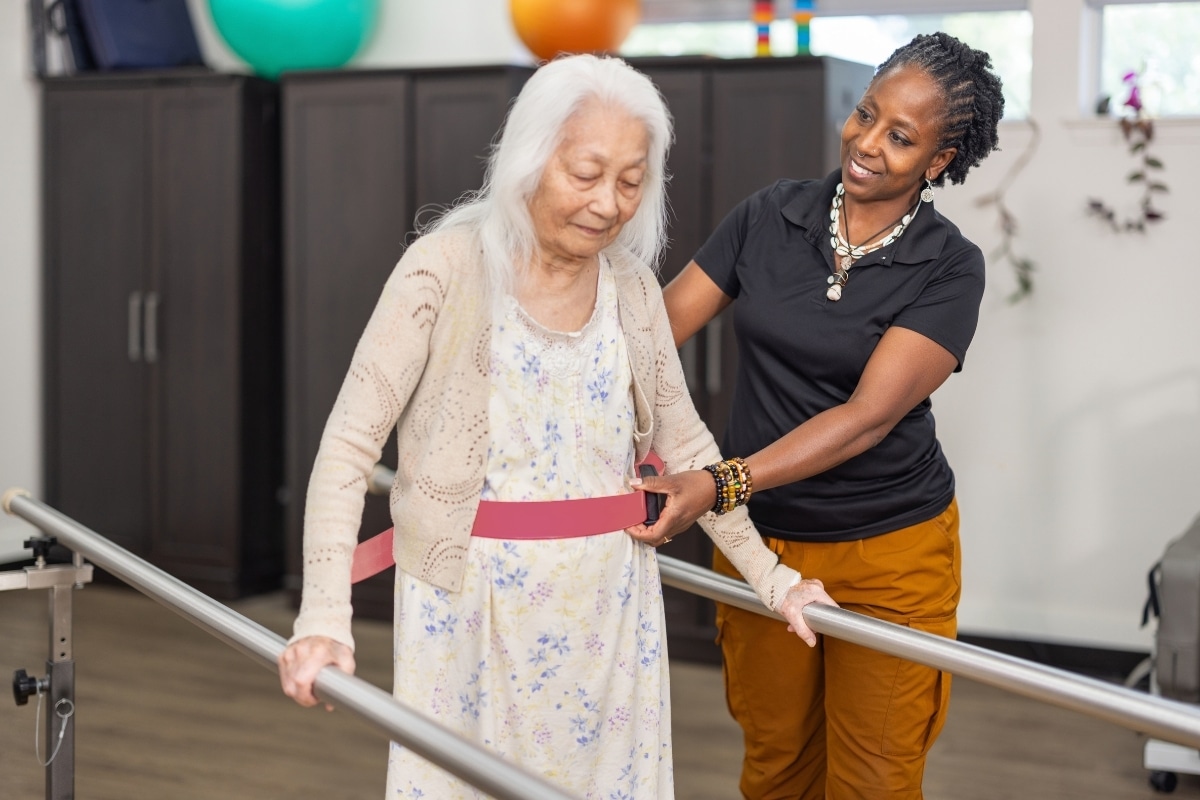 Staff member supporting a resident as she walks between parallel bars.